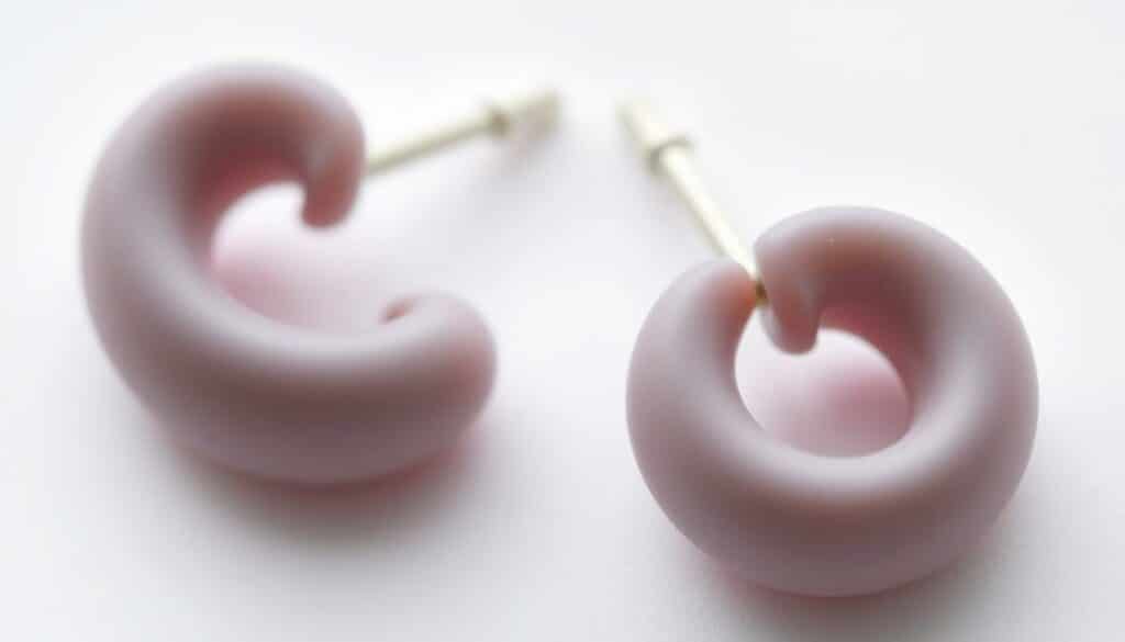 Detailed close-up photograph of a pair of plastic or silicone earrings, shot against a plain white background with soft, even lighting from above. The earrings are simple, contemporary in design, and appear smooth, lightweight, and hypoallergenic. The focus is sharp, highlighting the intricate textures and subtle reflections on the earring surfaces. The image conveys a sense of purity, cleanliness, and safety, reflecting the idea of an allergen-free accessory suitable for sensitive skin.